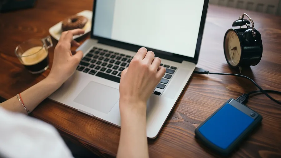 Hände einer Frau auf der Tastatur eines MacBooks, daneben eine externe blaue Festplatte am Kabel und ein Wecker auf dem Holztisch