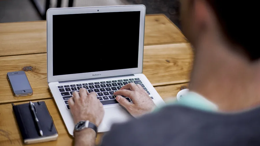 Mann von hinten tippt auf einem aufgeklappten MacBook Air, daneben liegen ein Smartphone und ein Notizbuch mit Stift auf einem Holztisch