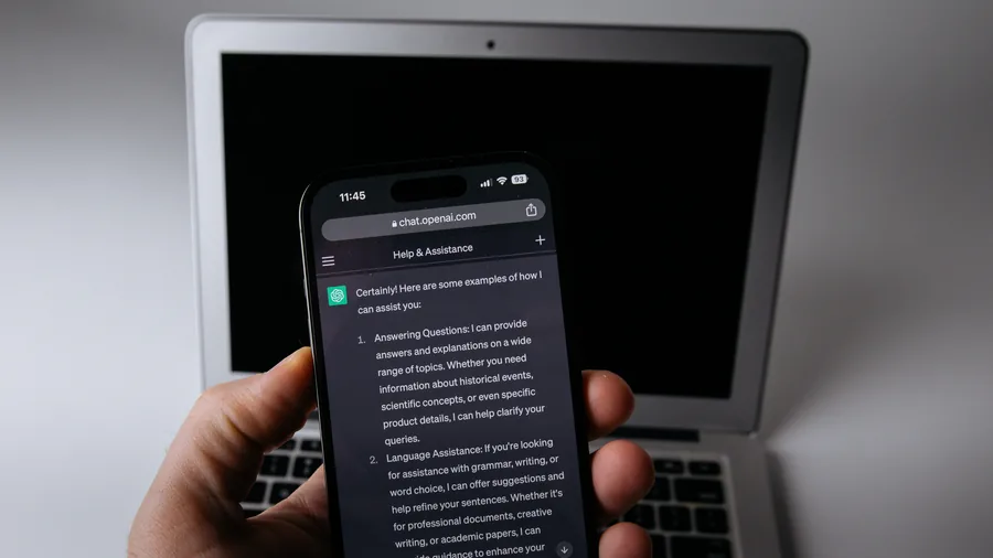 Eine Hand hält ein Smartphone mit geöffnetem ChatGPT vor einem aufgeklappten Laptop