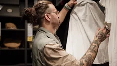Mann mit Brille und Haarknoten hält in einem Geschäft ein weißes Hemd von der Kleiderstange hoch und sieht es sich an
