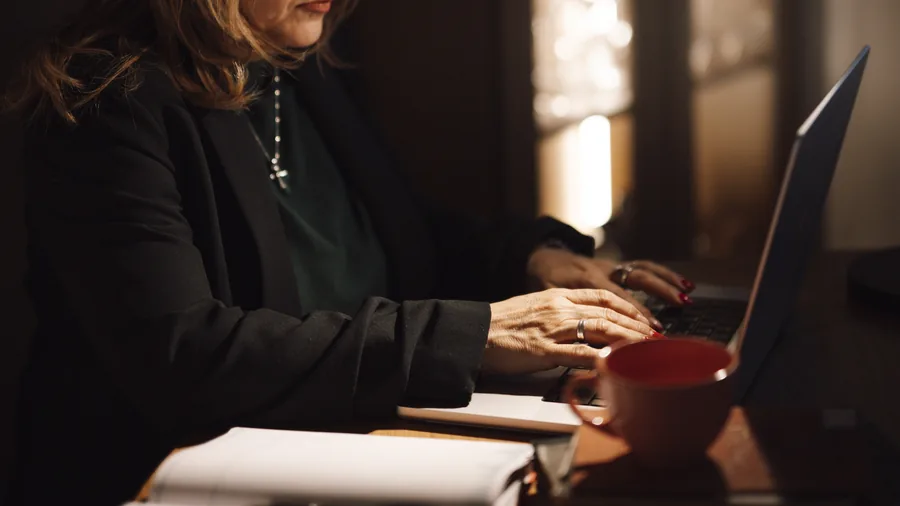 Eine Frau im Blazer tippt am Abend an einem Laptop, daneben eine rote Tasse und ein aufgeschlagenes Notizbuch
