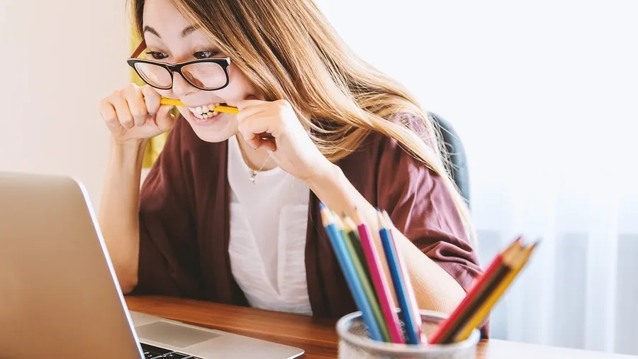 Junge Frau mit Brille beißt vor einem aufgeklappten Laptop auf einen Bleistift, vorne ein Becher mit Buntstiften