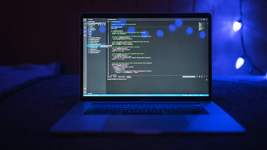 Aufgeklappter Laptop mit Programmcode auf dem Bildschirm in einem dunklen Raum mit blauem Licht