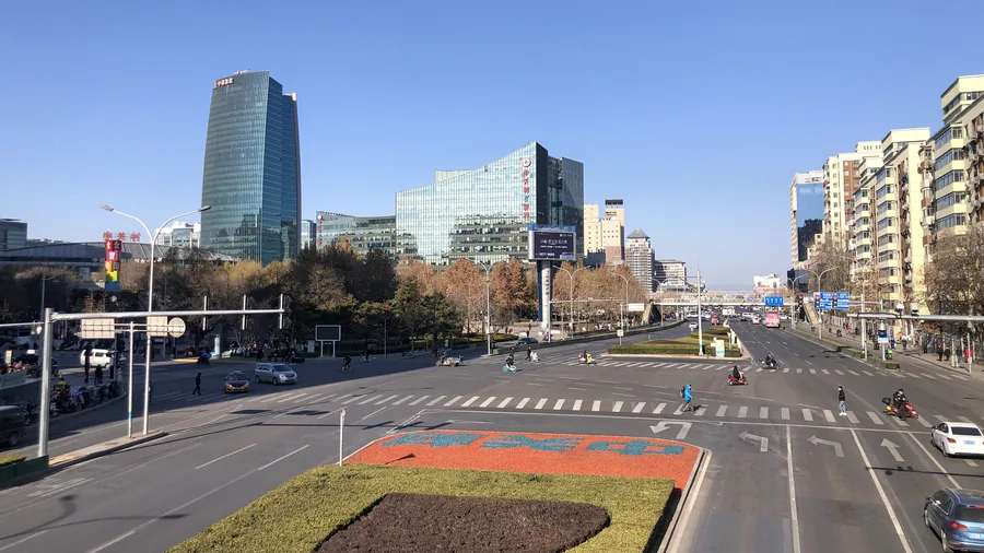 Breite Kreuzung im Pekinger Technologieviertel Zhongguancun mit Glasbürotürmen, Radfahrern und einem roten Schriftzug im Blumenbeet