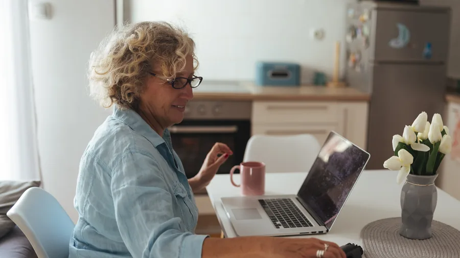 Ältere Frau mit Brille sitzt in ihrer Küche am Esstisch und arbeitet am Laptop, daneben Kaffeetasse und Blumen