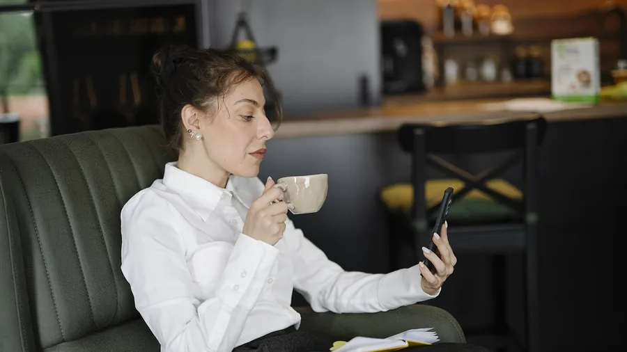 Frau in weißer Bluse sitzt in einem grünen Sessel, hält eine Tasse und schaut auf ihr Smartphone, auf dem Schoß liegt ein Notizbuch