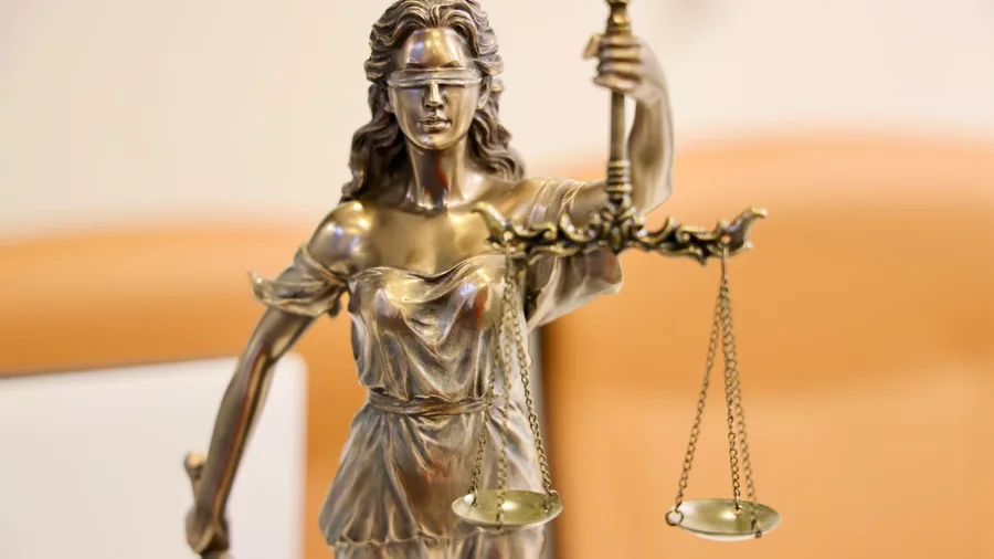 Eine Bronzefigur der Justitia mit verbundenen Augen hält eine Waage in die Höhe, der Hintergrund ist unscharf und orange