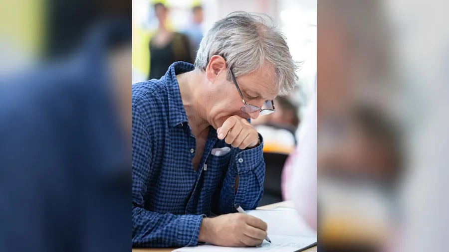 Axel Scheffler mit grauem Haar, Brille und blau kariertem Hemd stützt das Kinn in die Hand und zeichnet mit einem Stift auf ein Blatt Papier