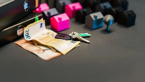 Kleine Kunststoffhäuser, Euro-Scheine und ein Schlüsselbund auf einem dunklen Tisch, links daneben zwei Ordner