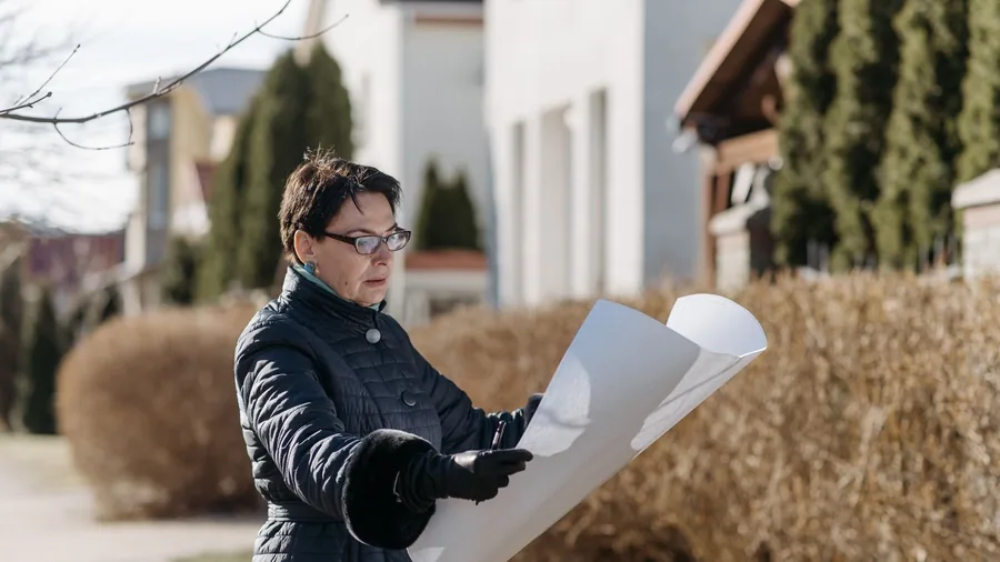 Frau mit Brille und Steppjacke hält in einer Wohnsiedlung einen großen Plan in der Hand