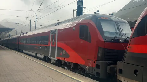 Roter ÖBB-Railjet der 2. Generation am Bahnsteig im Bahnhof Landeck-Zams, im Hintergrund Berge und bewölkter Himmel
