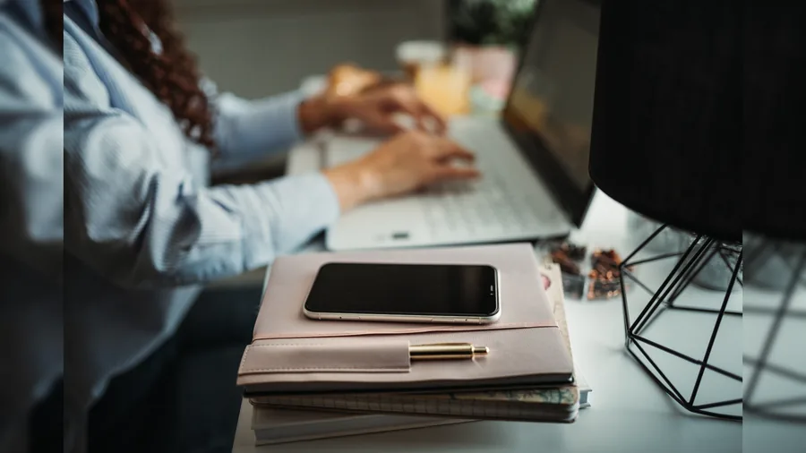 Person arbeitet konzentriert an einem Laptop, daneben liegen Smartphone und Notizbuch auf dem Schreibtisch