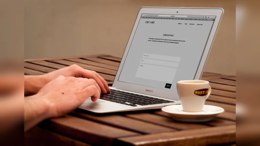 Hände tippen auf einem Laptop, daneben steht eine Tasse Kaffee auf einem Holztisch