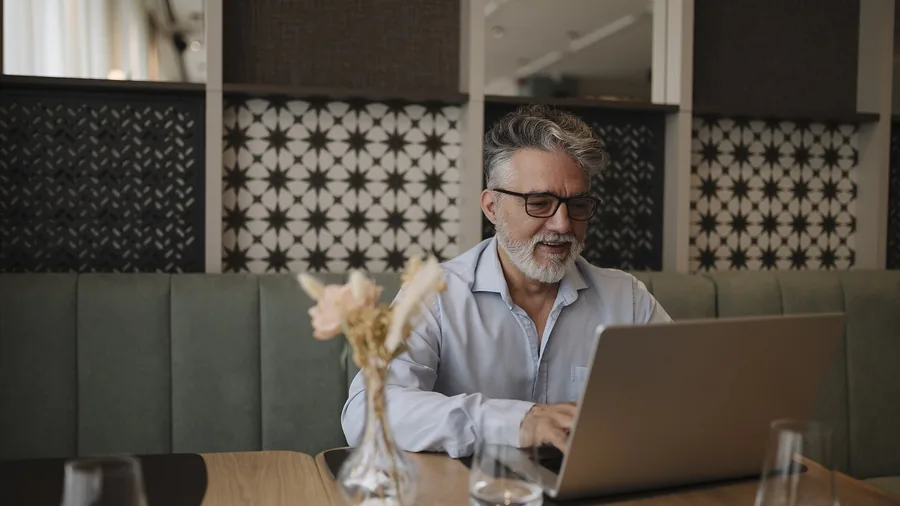 Mann mit grauem Bart und Brille tippt lächelnd auf einem Laptop an einem Holztisch in einem Café