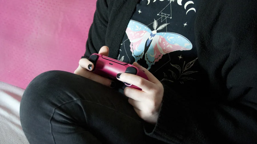 Person in schwarzem Pullover sitzt mit überkreuzten Beinen und hält einen roten PS5-Controller in beiden Händen