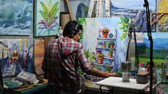 Maler mit Kopfhörern steht im Atelier vor einer bunten Leinwand mit Blumentöpfen auf einer Leiter, rundherum hängen seine Bilder