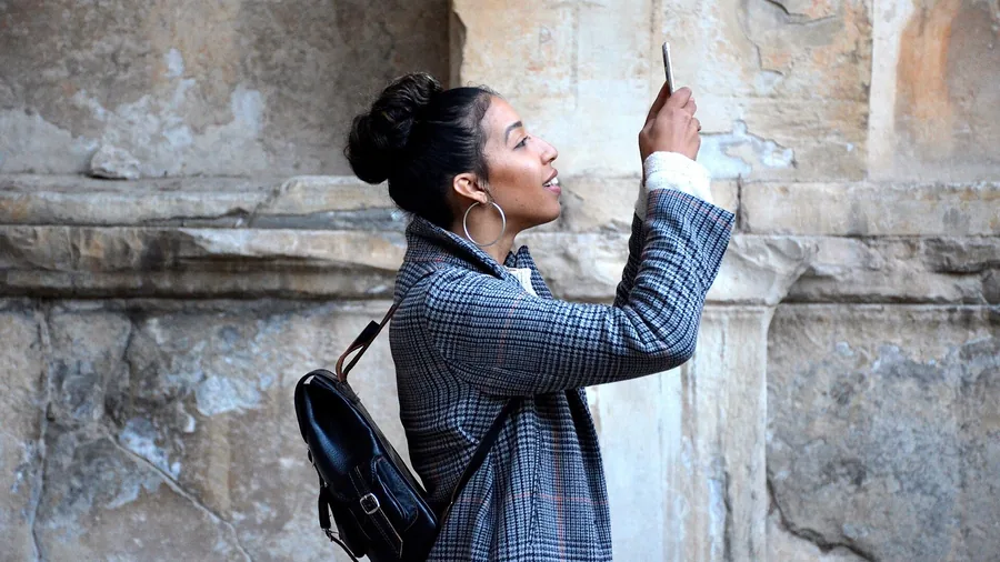 Frau mit Rucksack hält ihr Handy hoch und fotografiert eine alte Steinmauer